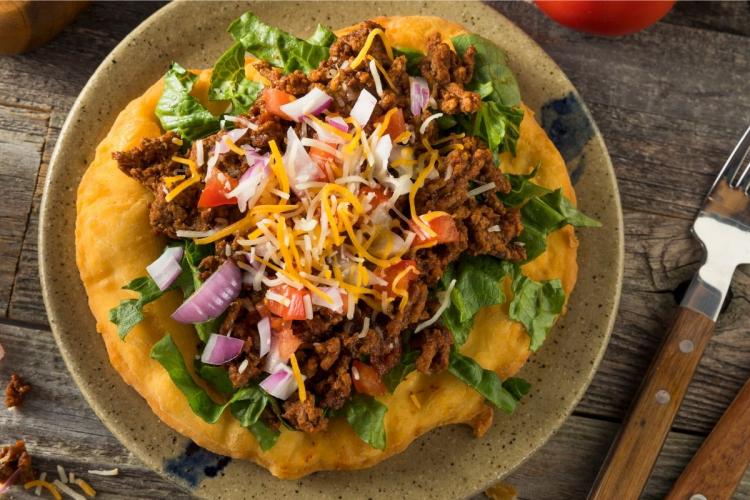 A mouthwatering New Mexican taco indio filled with slow-cooked meat, fresh lettuce, diced tomatoes, chopped red onion and shredded cheese, served on a ceramic plate.