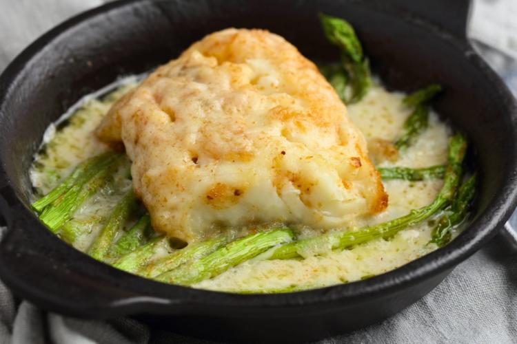 A fillet of baked haddock in cream sauce, accompanied with asparagus, in an individual serving plate.